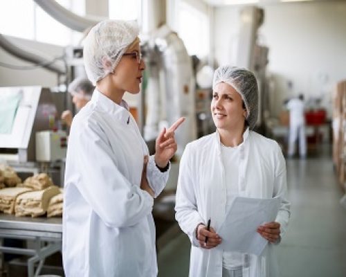 Two female workers discussing while standing in food factory. One of them holding paperwork and pen while other one speaking.